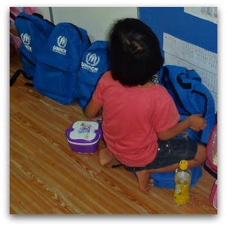 Little girl in pink T-shirt crouches near blue UNHCR backbacks