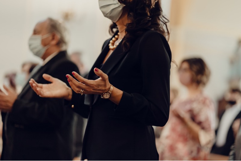 Main image: Woman in church wearing a face mask and praying with her hands outstretched. Other pray in the background. (Gabriella Clare Marino/Unsplash)
