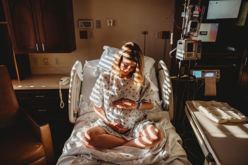 Pregnant woman touching stomach while sitting on bed in hospital (Credit: Cavan Images)